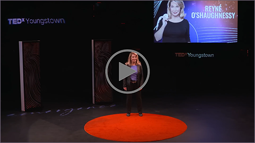 A woman stands on a red circular carpet delivering a talk at a TEDxYoungstown event. A screen behind her displays her name and photo.