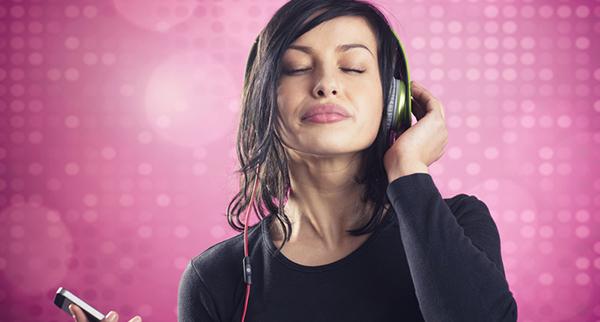 A person with closed eyes listens to music on headphones, holding a phone. The background is pink with circular patterns.