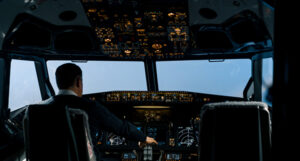 Airplane cockpit with a pilot seated at the controls, surrounded by illuminated instrument panels, against a view of the sky.