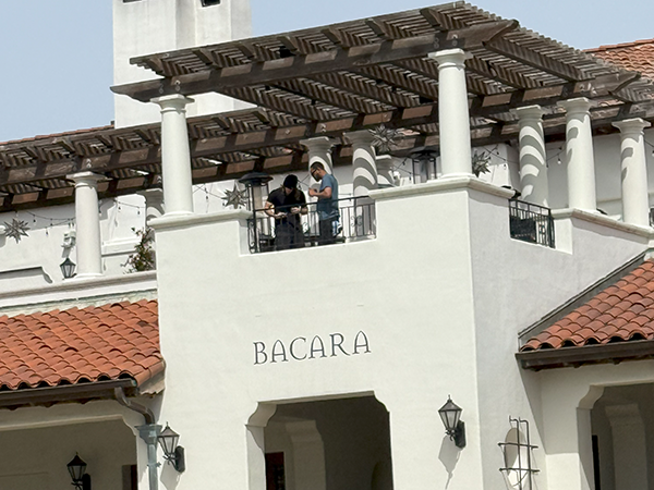 Two people stand on a balcony of a white building with a red-tiled roof and columns, under a pergola, with the word "BACARA" on the wall below them.