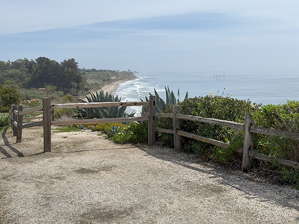 Wooden fence overlooking a coastal cliff with waves, greenery, and distant hills under a cloudy sky.