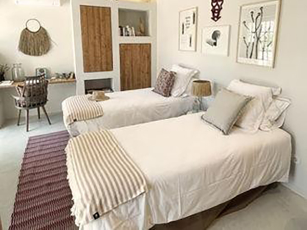 Two neatly made twin beds in a bedroom with a desk and chair by the window. Walls feature minimalist decor and framed art. A striped rug lies on the floor.