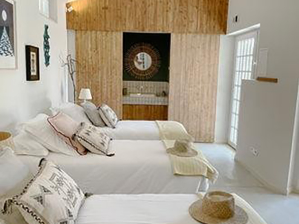 A bedroom with three neatly made beds, decorative pillows, and straw hats. Wooden paneling on one wall, framed art, and an open doorway leading to a sink area. Natural light filters through a window.