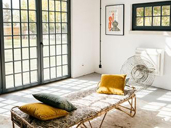 A sunlit room with a rattan daybed, green and yellow cushions, and modern grid-patterned windows. A piece of abstract art and a wire sculpture decorate the walls.
