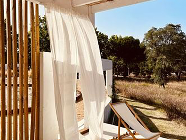 A white curtain blows in the wind on a patio with a wooden chair nearby, overlooking a grassy area with trees under a clear sky.
