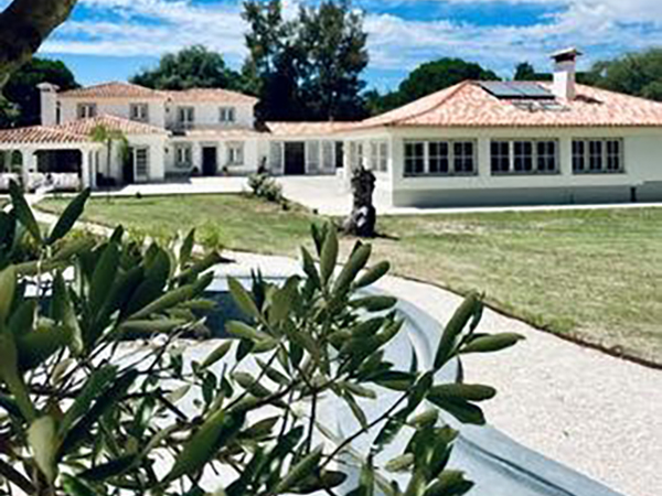 Large white villa with a terracotta roof, surrounded by greenery and a spacious lawn, under a partly cloudy sky.
