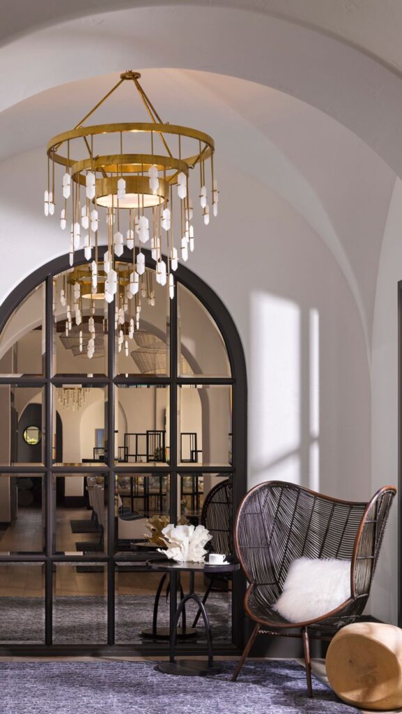 Elegant room with a large arched window, gold chandelier, black wireframe chair with white cushion, a small round wooden table, and a gray rug.