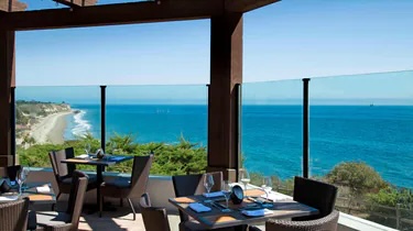Restaurant patio with dining tables and chairs overlooking a coastline, under a partly cloudy blue sky.