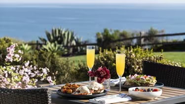 Outdoor table with two plates of breakfast, salads, and two glasses of orange juice. Ocean and garden view in the background.