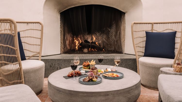 A cozy indoor setting with a lit fireplace. Plates of food, including steak and pasta, are on a round table surrounded by cushioned chairs. Two glasses of wine accompany the meal.