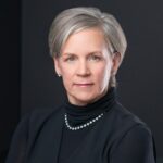A middle-aged woman with short gray hair, wearing a black top and pearl necklace, poses against a dark background.