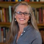 A woman with long, light brown hair and glasses smiles at the camera, standing in front of shelves filled with books.