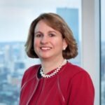 A woman with short brown hair, wearing a red jacket and pearl necklace, smiles in front of a window with a cityscape in the background.