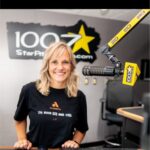 A woman with blonde hair stands smiling in a radio studio next to a microphone branded with the 100.7 Star logo.
