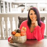 A woman in a bright pink blouse sits at a table, smiling, and holding a bowl filled with assorted fruits.