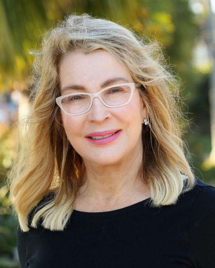 Woman with glasses and shoulder-length blonde hair smiling outdoors in a black top.