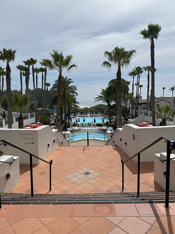 View of a sunny resort pool area with lounge chairs, surrounded by palm trees and hills in the background.
