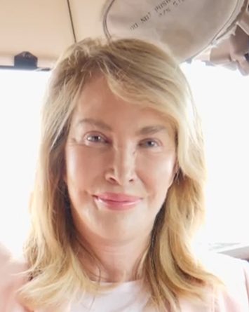 A person with shoulder-length blonde hair is smiling slightly while sitting inside a vehicle with light coming through the window behind them.