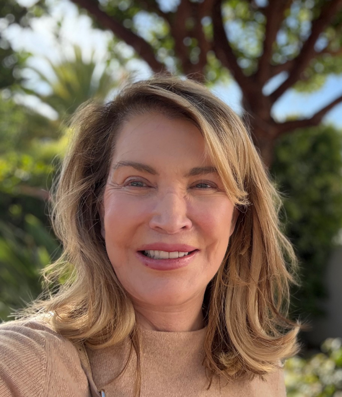 Smiling woman with shoulder-length hair takes a selfie outdoors, with trees and greenery in the background.