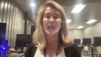 A woman with blonde hair is speaking in an office or computer lab with several monitors and desks visible in the background.